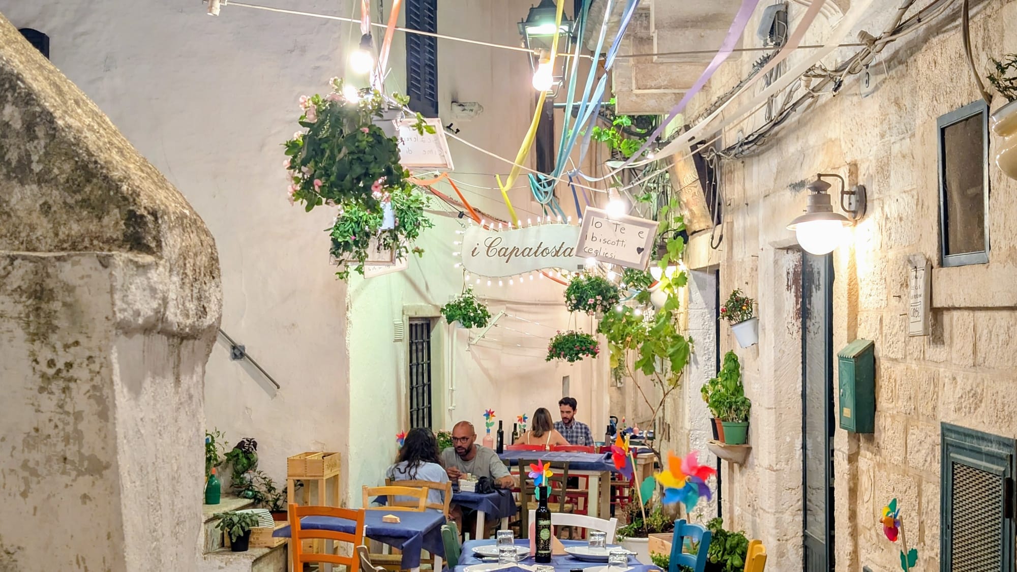 Abends in einer Gasse von Ceglie Messapica: gedeckte Tische mitten auf dem Kopfsteinpflaster, bunte Stühle, Wimpel und Lichterketten zwischen den Kalksteinhäusern